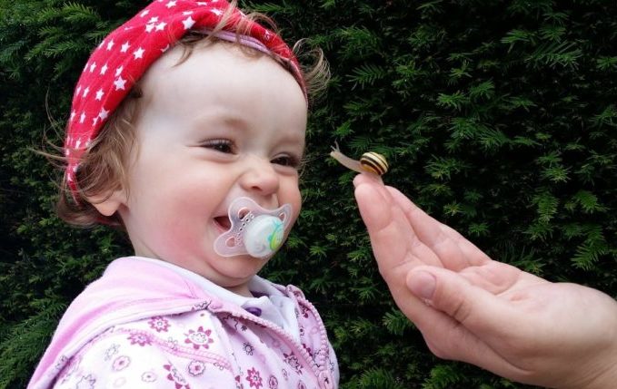 Kind mit Schnuller im Mund lacht wegen Schnecke auf erwachsener Hand