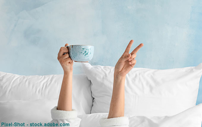 Frau liegt im Bett und hält Tasse in der Hand und zeigt Peace Zeichen.jpg